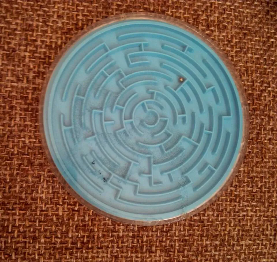 Overhead view of a round blue labyrinth puzzle toy with a metal ball on a woven brown textured surface