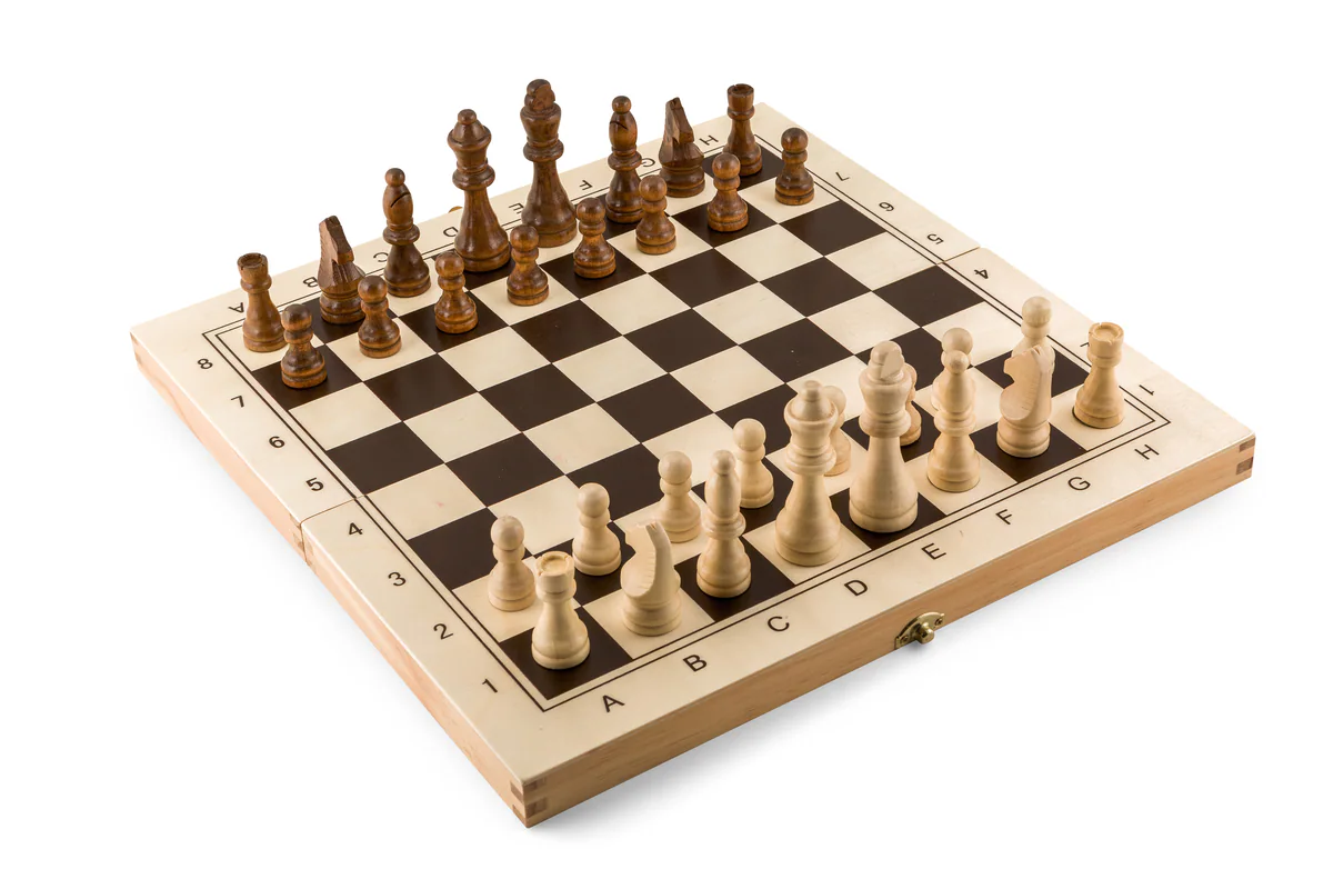 A wooden chess set with light and dark pieces arranged at the start of a game on a folding board, shot against a plain warm background